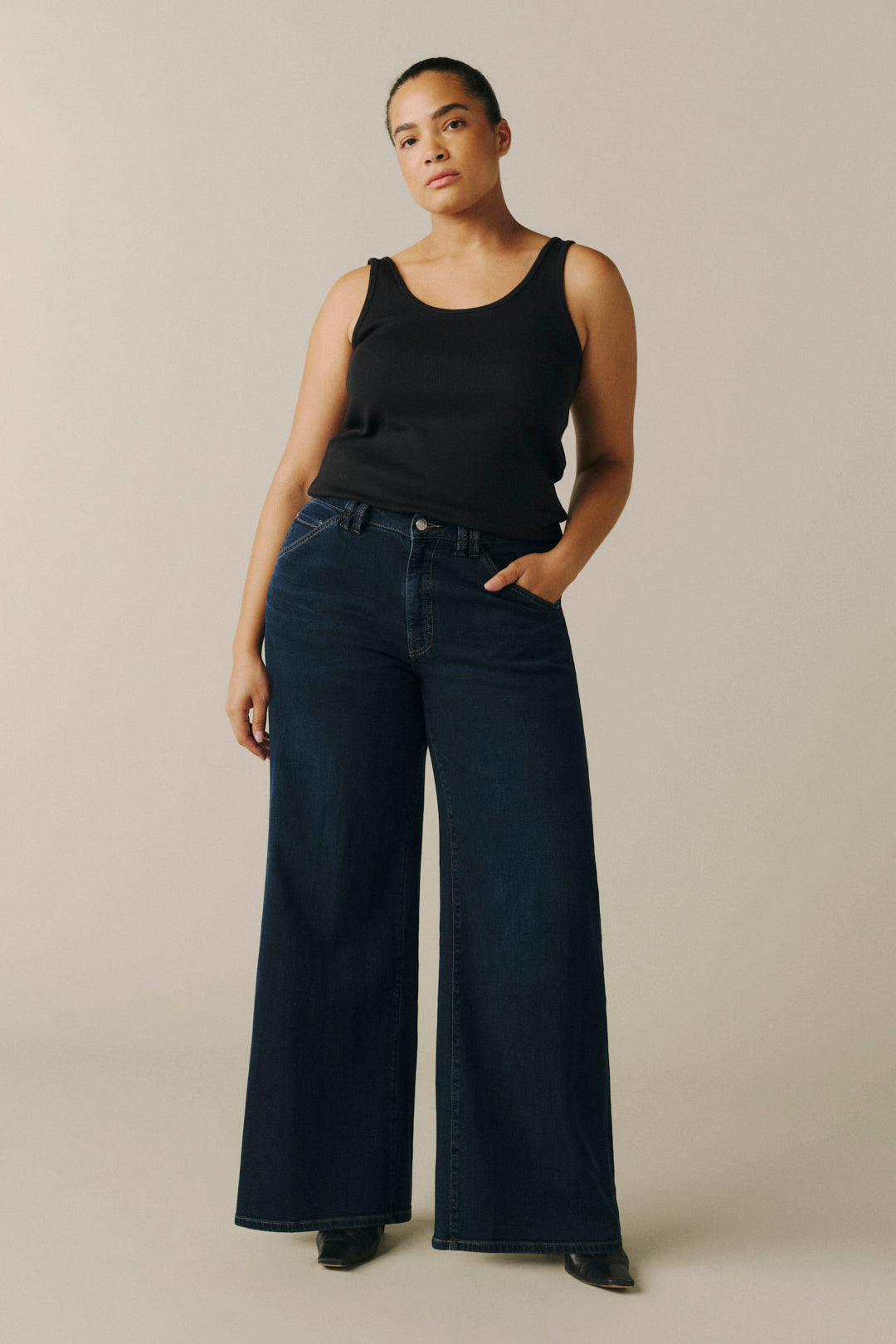 A model wearing a black sleeveless tank top and dark wide-leg denim jeans, standing against a plain beige studio background.