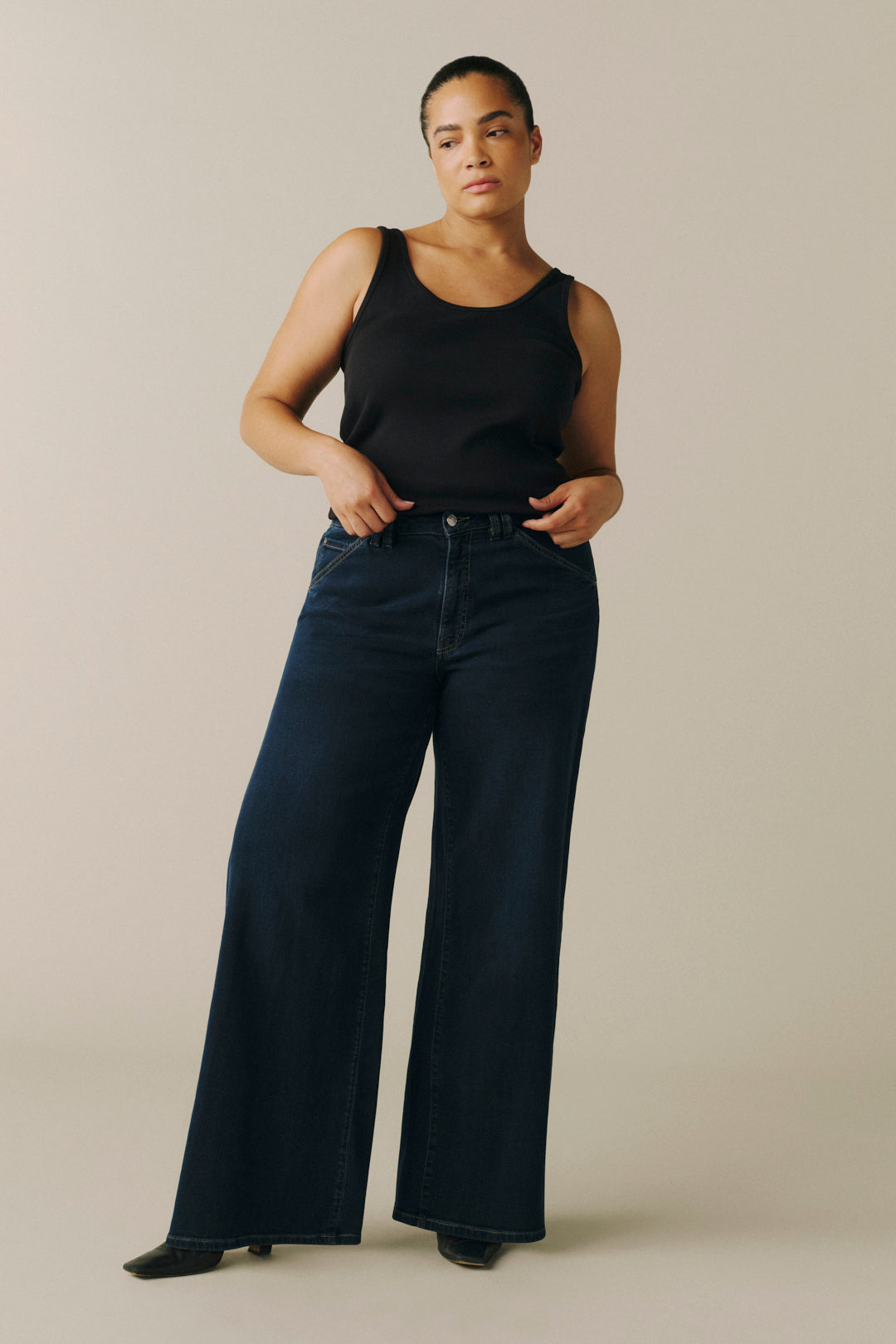 A woman modeling dark blue wide-leg denim jeans and a black tank top against a plain beige studio background.