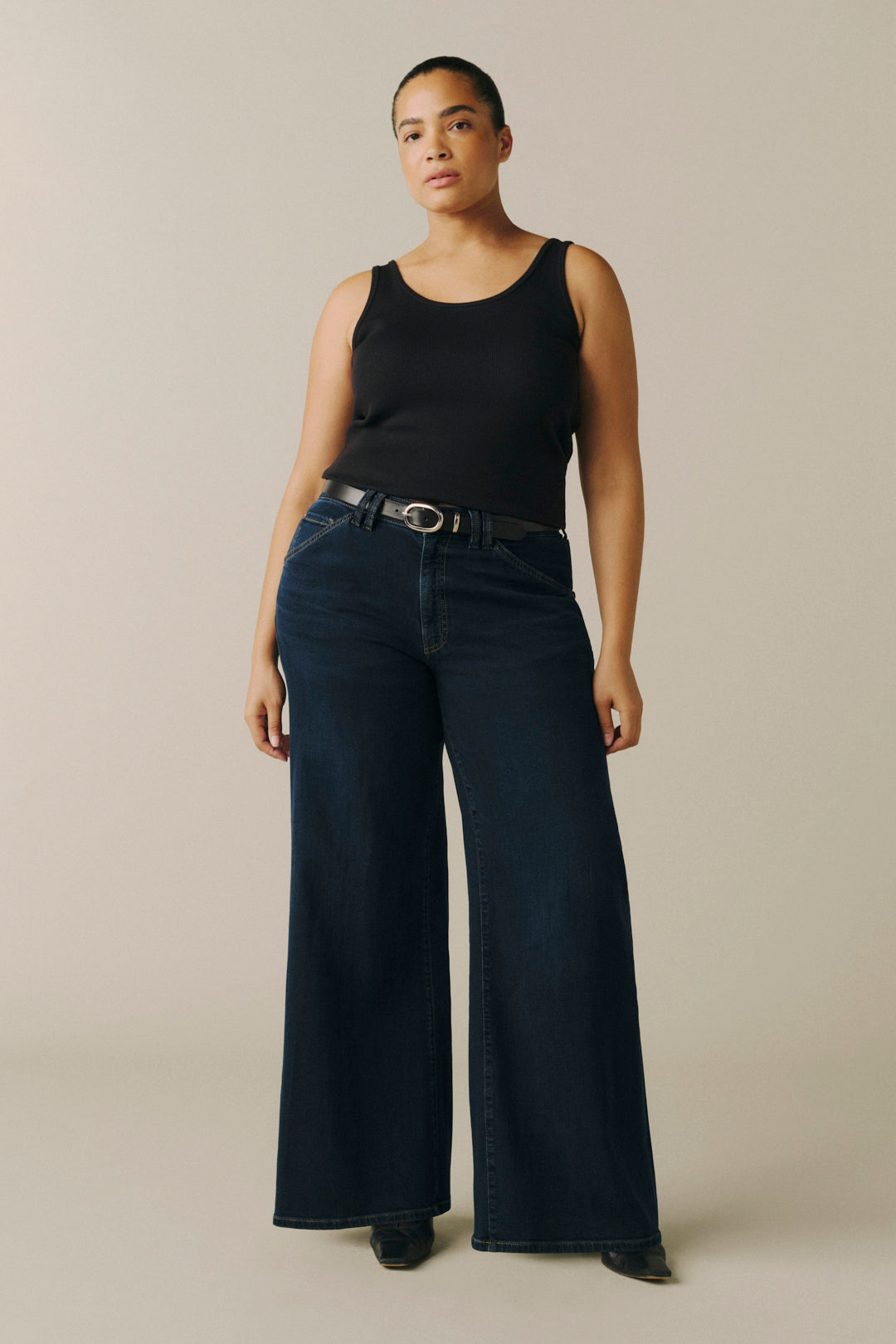 A model stands against a plain beige studio background wearing a black sleeveless top, dark wide-leg denim jeans with a black belt, and black shoes.