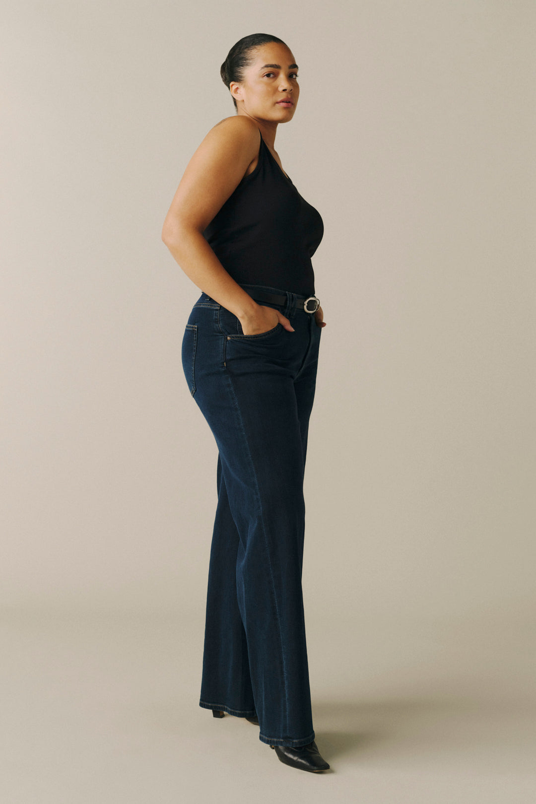 A woman modeling dark blue flared jeans with a silver-tone belt, paired with a black sleeveless top, standing in profile against a solid beige studio background.