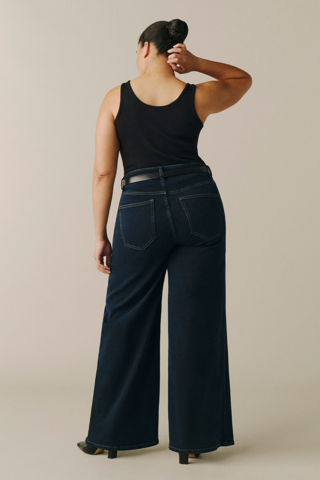 A woman shown from the back wearing dark wide-leg jeans with a thin black belt, a black sleeveless top, and black heels, standing against a plain light beige studio background.