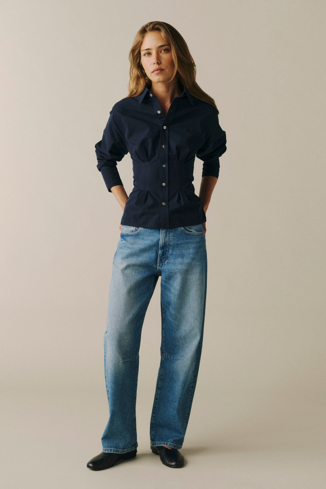 A woman modeling a dark navy blue long-sleeve button-up shirt with a peplum waist and front pockets, paired with blue jeans and black shoes, against a solid beige background.