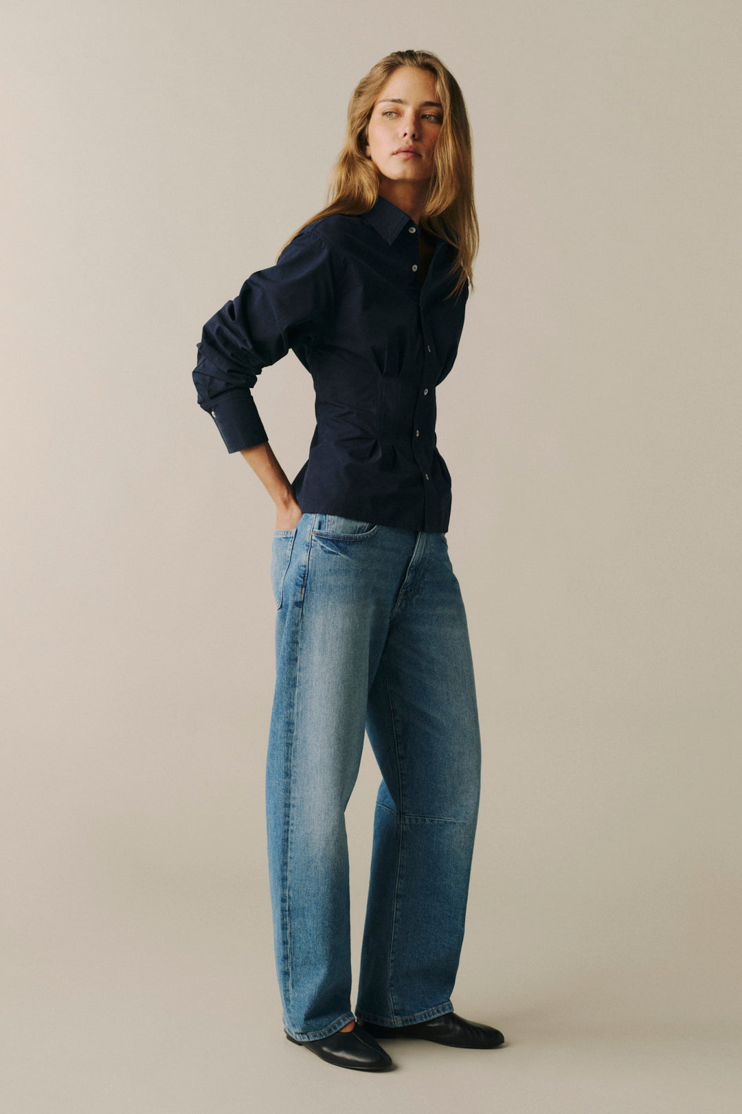 A woman modeling a dark navy blue button-up shirt and wide-leg blue jeans against a solid light beige studio background.