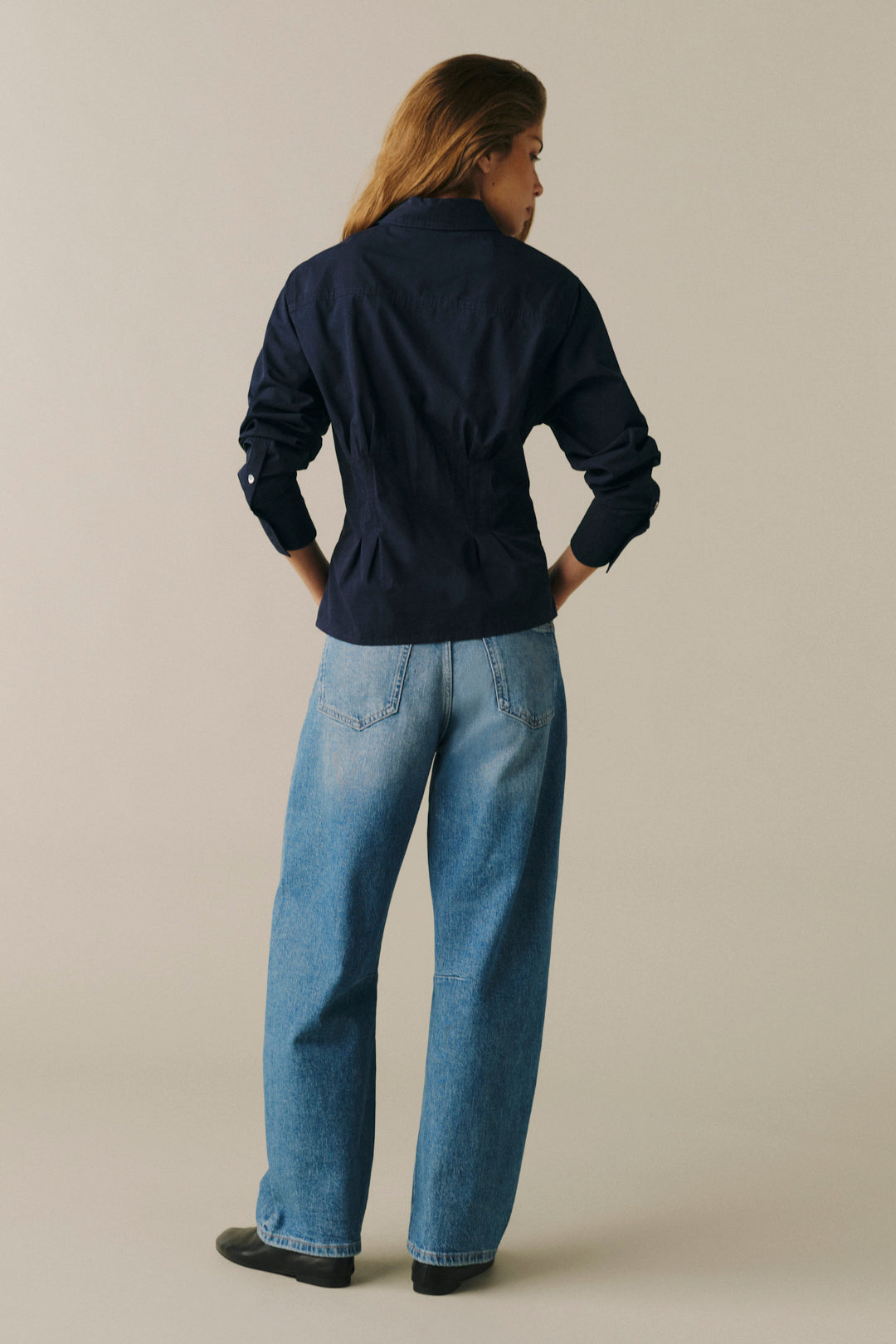A woman is shown from behind wearing a dark navy blue long-sleeve jacket and light blue wide-leg jeans, standing against a solid light beige studio background.