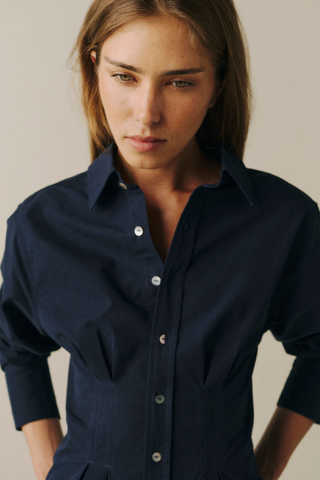 A woman wearing a dark navy blue long-sleeve button-up shirt, shown from the chest up against a plain light beige background.
