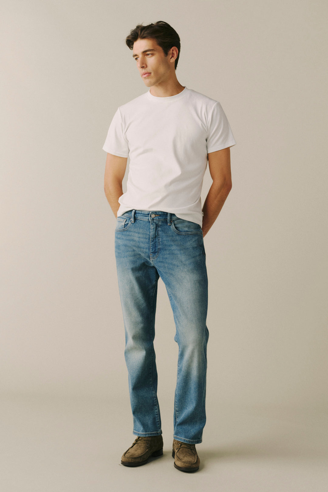 A man wearing a white crewneck t-shirt, blue straight-leg jeans, and brown suede shoes, standing against a neutral beige studio background.