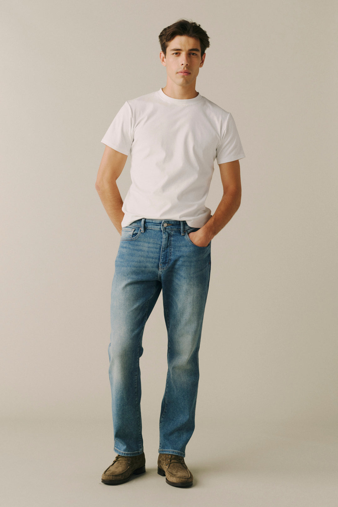 A man wearing a white crewneck t-shirt, blue straight-leg jeans, and brown suede shoes, standing against a solid light beige studio background.