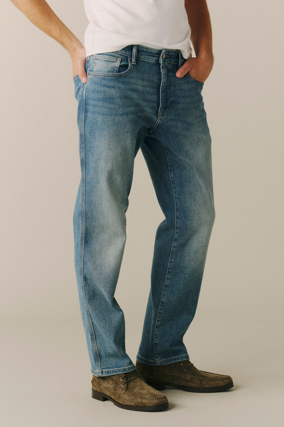 A person wearing blue denim jeans with a faded wash, paired with a white t-shirt and brown suede shoes, standing against a neutral beige studio background.
