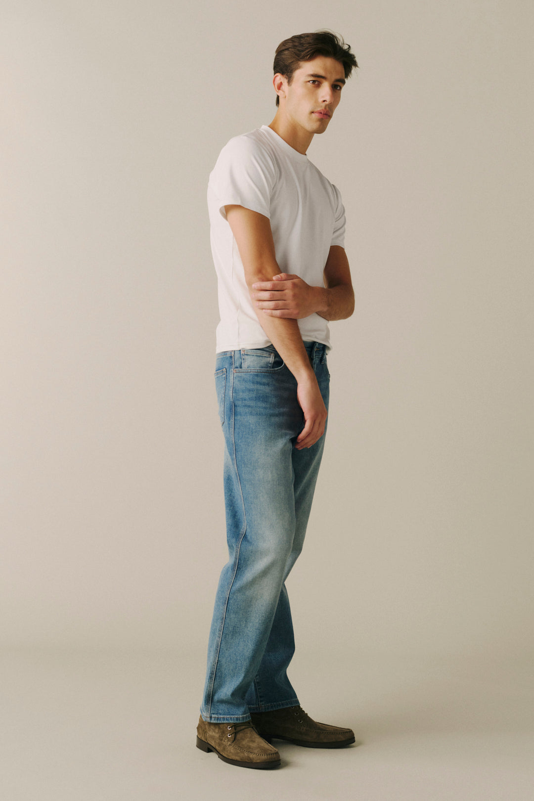 A man wearing a white crewneck t-shirt, blue jeans, and brown suede boots, standing in profile against a plain light beige studio background.