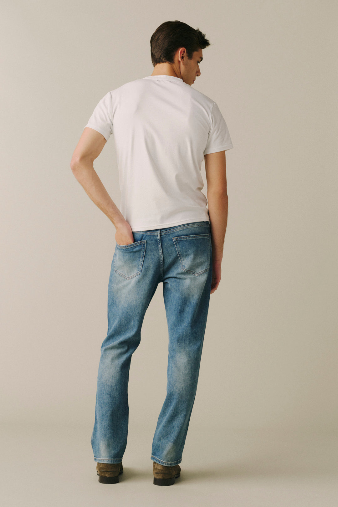 A man viewed from behind wearing a white t-shirt and light blue denim jeans, standing against a plain beige studio background.