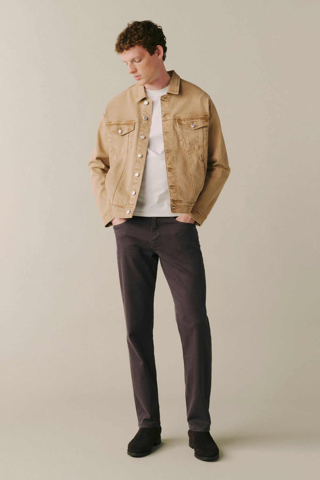 A man wearing a tan denim jacket over a white t-shirt, dark charcoal pants, and black shoes.