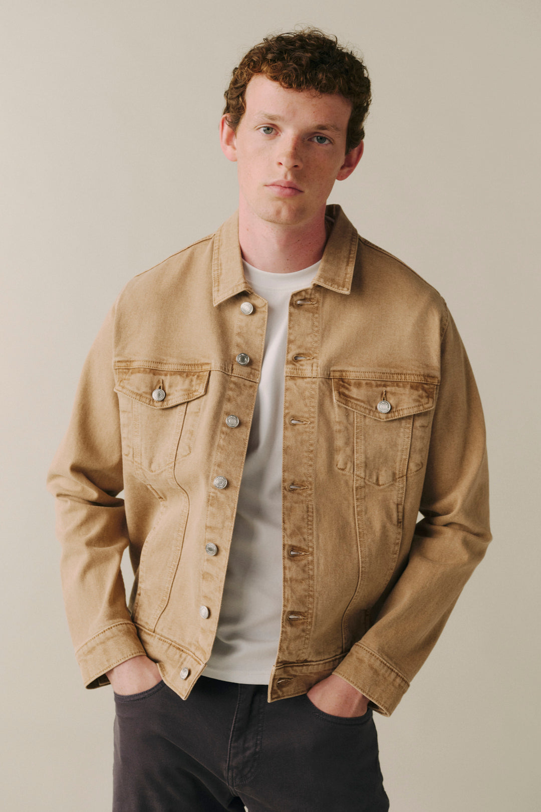 A man wearing a tan denim trucker jacket over a white t-shirt and dark gray pants, standing against a plain beige background.