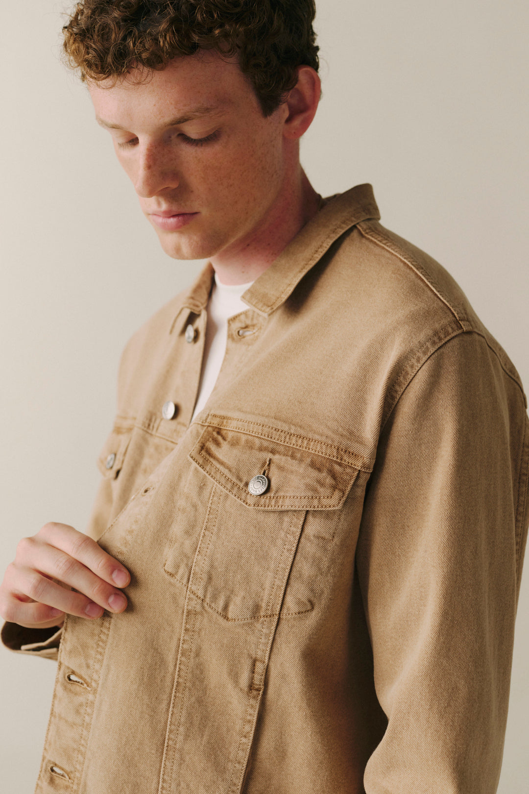 A man wearing a tan canvas utility jacket over a white t-shirt, shown in a close-up shot against a neutral beige background.