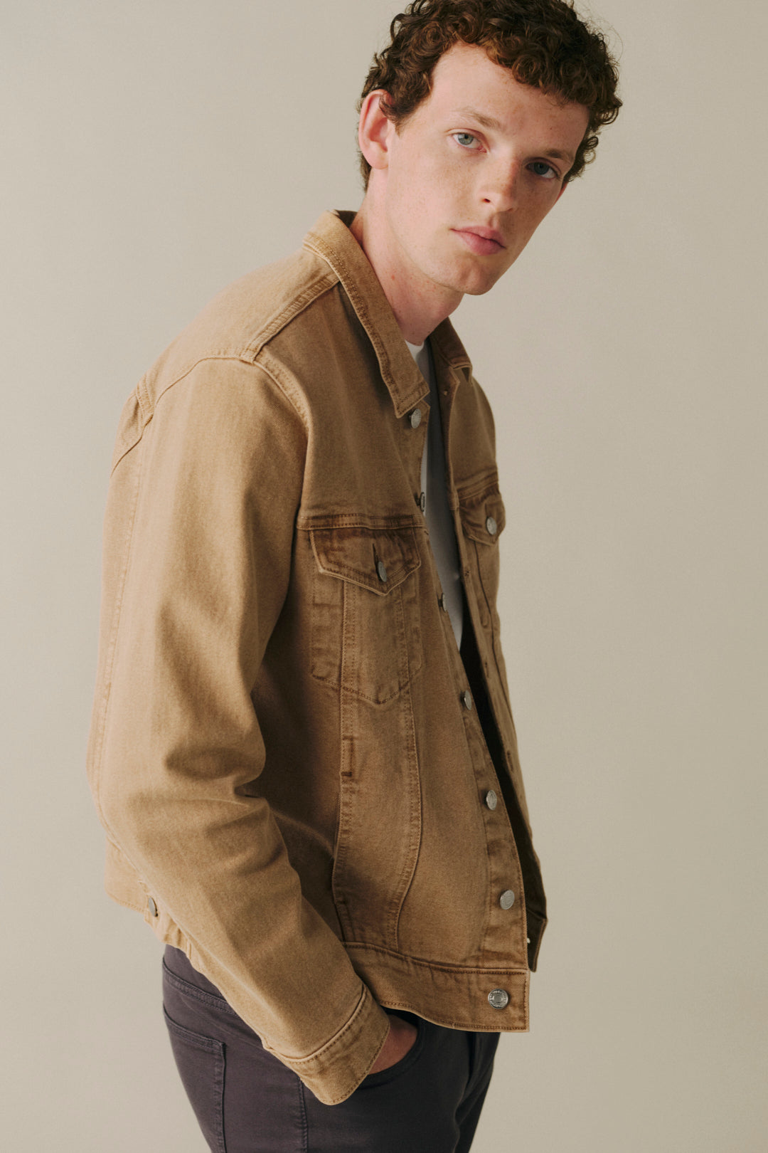 A man wearing a tan denim jacket over a dark shirt and dark pants, photographed against a plain light gray studio background.