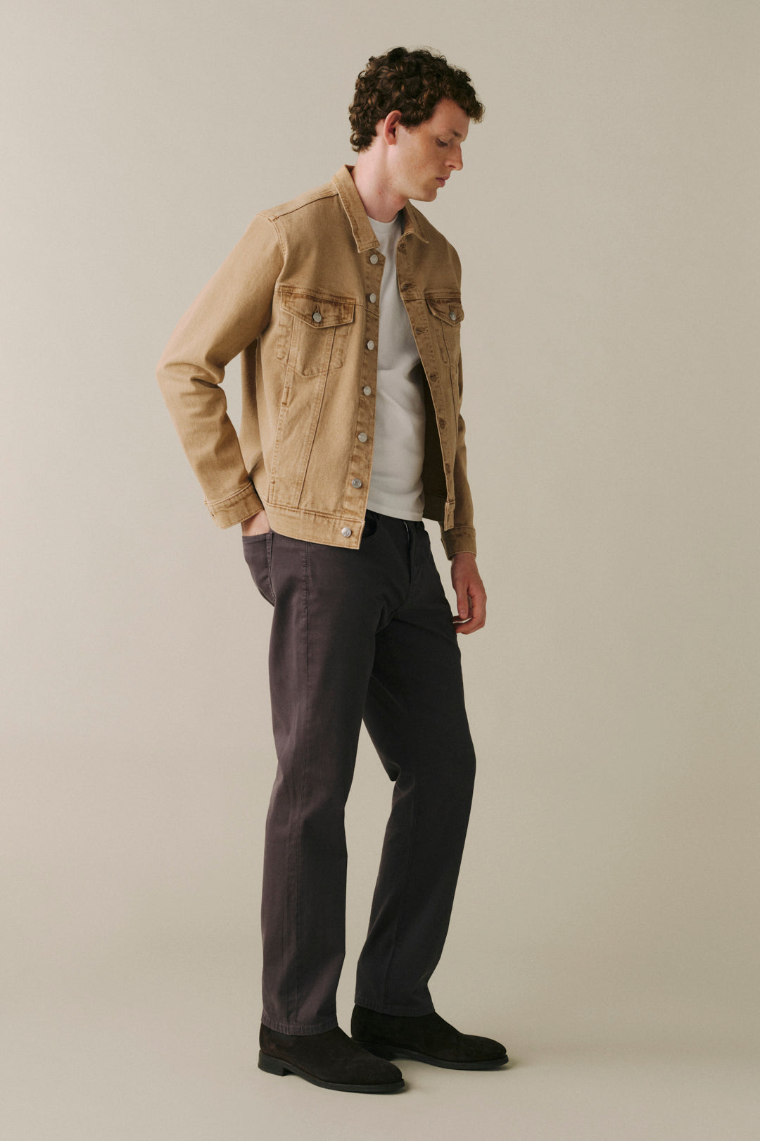 A man wearing a tan denim jacket over a white t-shirt, dark charcoal gray trousers, and black shoes.