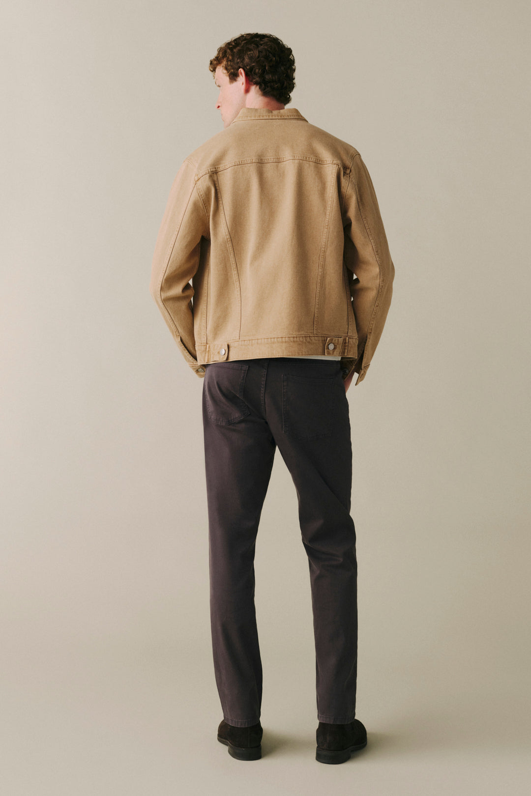 A man is shown from behind wearing a tan canvas utility jacket and dark charcoal gray trousers.