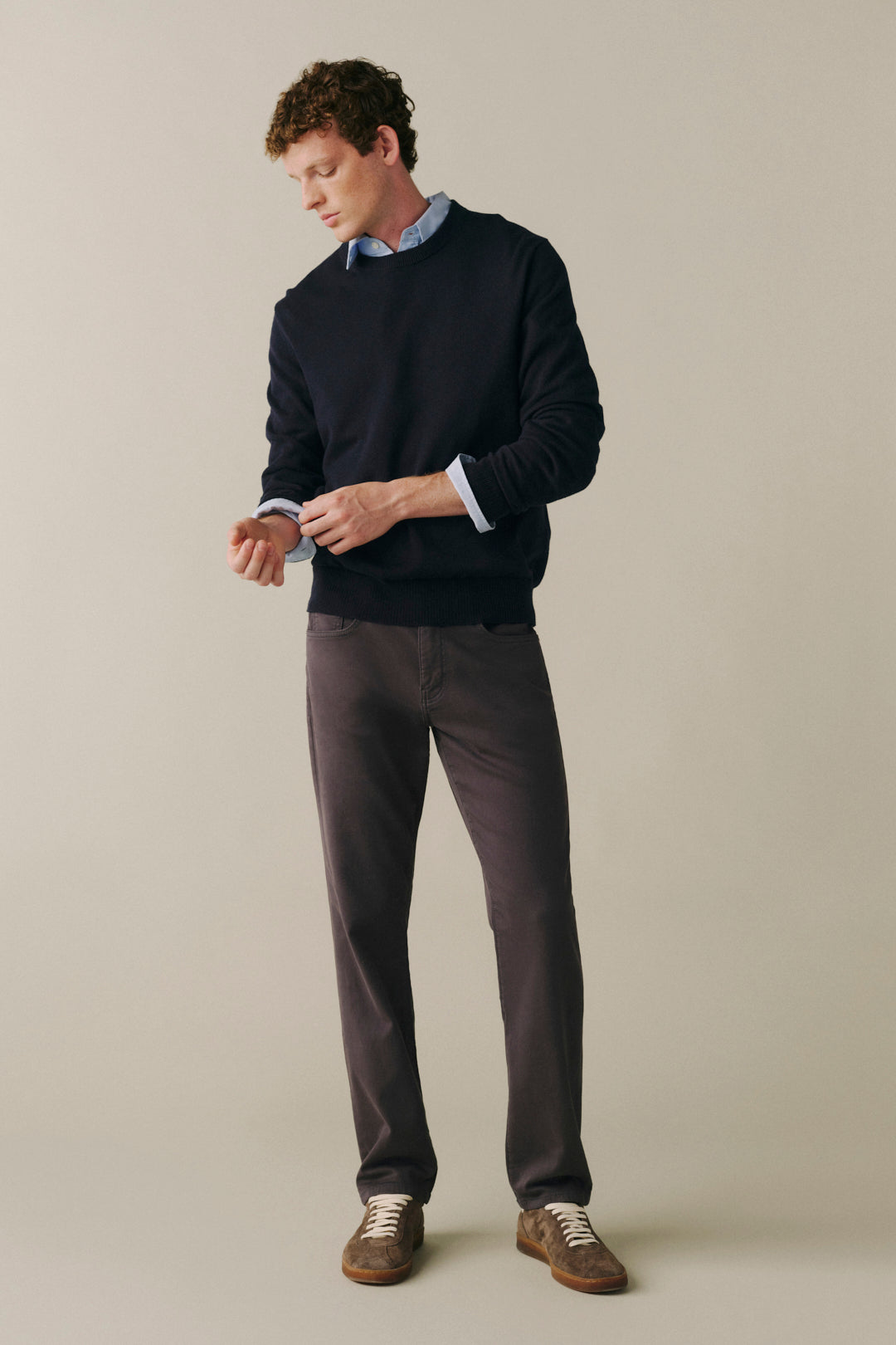 A man wearing a dark navy crewneck sweater, a light blue collared shirt, dark gray trousers, and brown suede shoes, standing against a solid beige background.