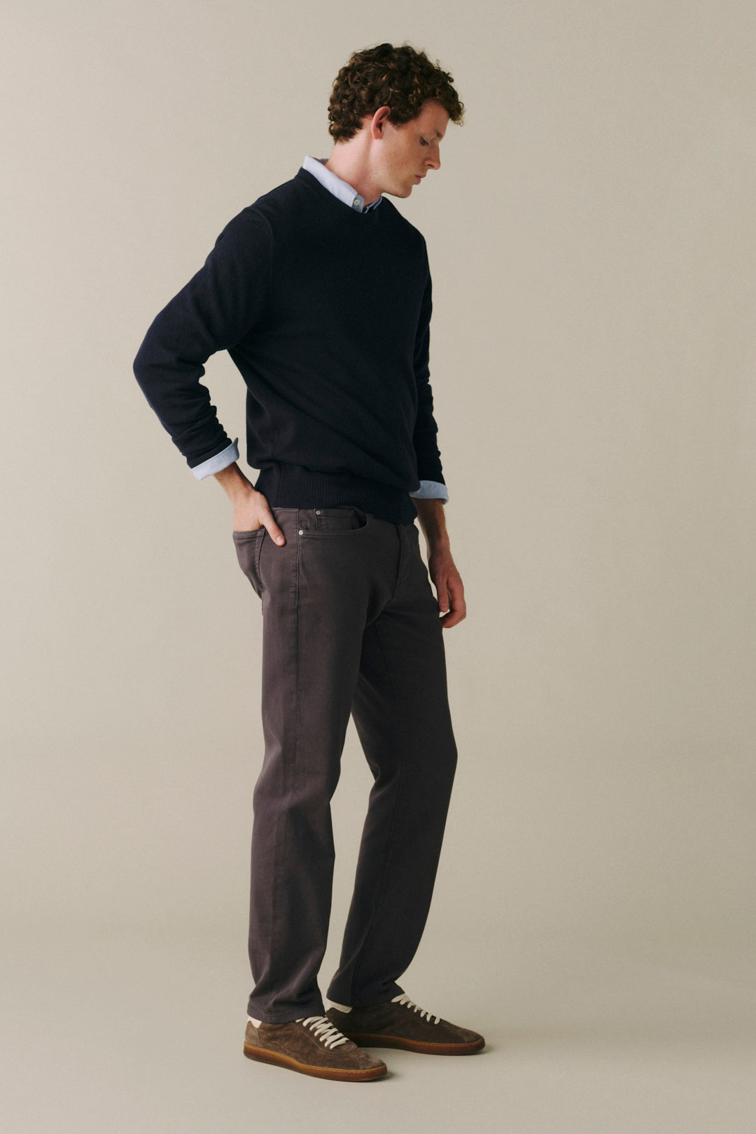 A man wearing a dark navy crewneck sweater, dark charcoal gray trousers, and brown suede shoes, standing in profile against a solid light beige background.
