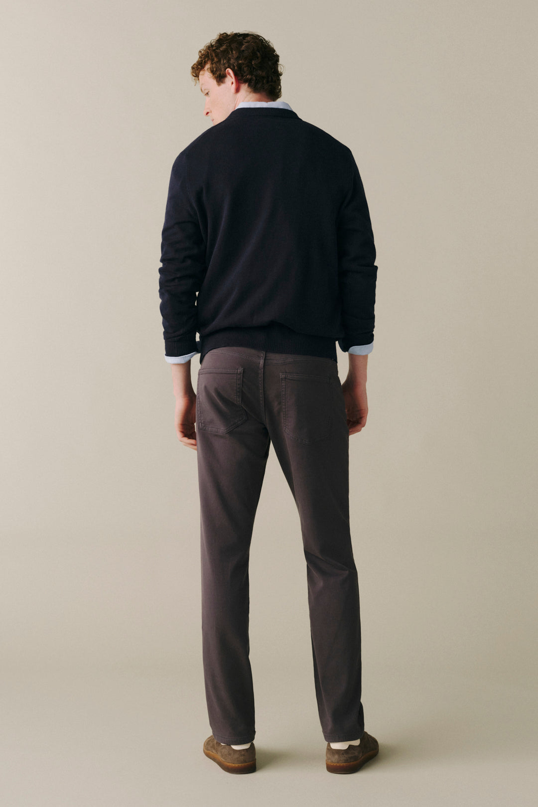 A man is shown from behind wearing a dark navy blue sweater, charcoal gray trousers, and brown shoes, standing against a solid light beige background.