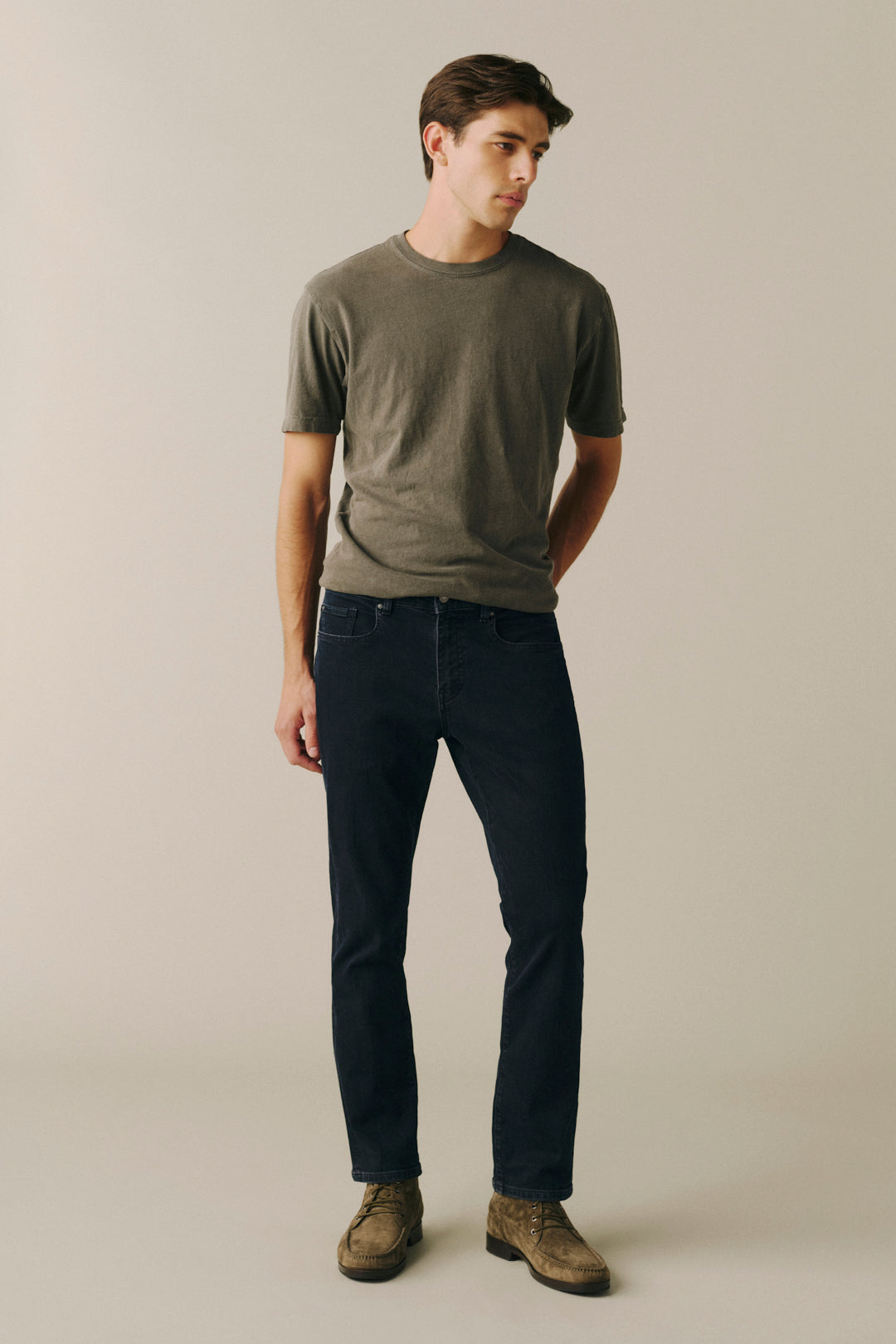 A man modeling a dark olive green crewneck t-shirt and dark navy blue jeans, paired with brown suede boots, against a plain beige studio background.