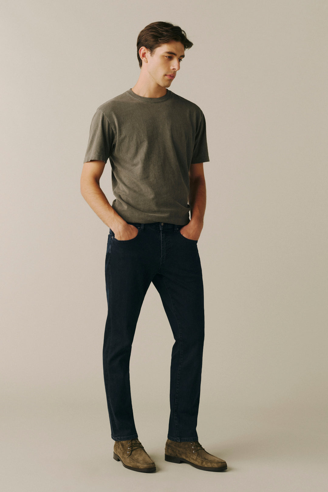 A man modeling a dark olive green t-shirt and dark wash jeans, standing against a solid light beige background.