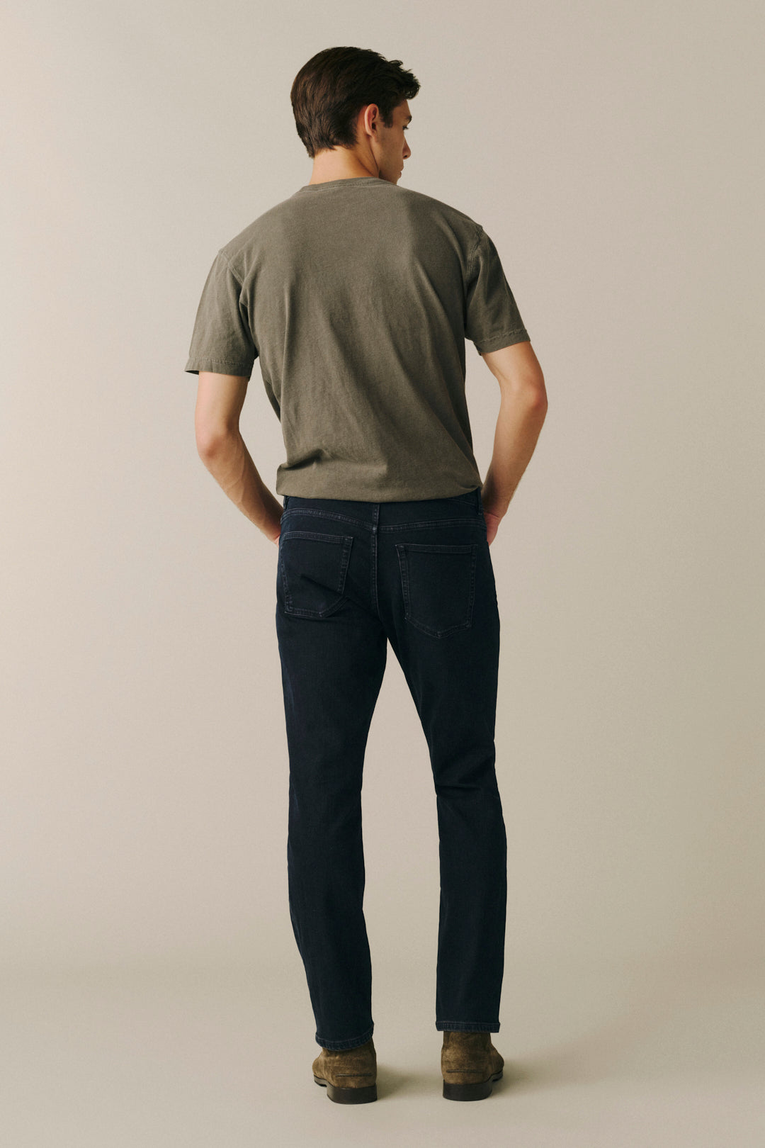 A man viewed from behind wearing a muted olive green t-shirt, dark navy blue jeans, and brown boots, standing against a plain light beige studio background.