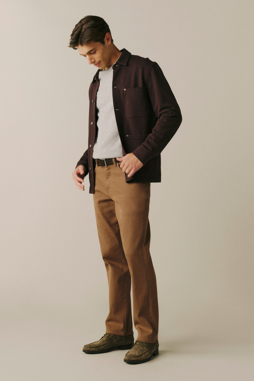 A man wearing a dark brown overshirt over a white t-shirt, brown trousers, and brown shoes, standing against a plain light beige studio background.