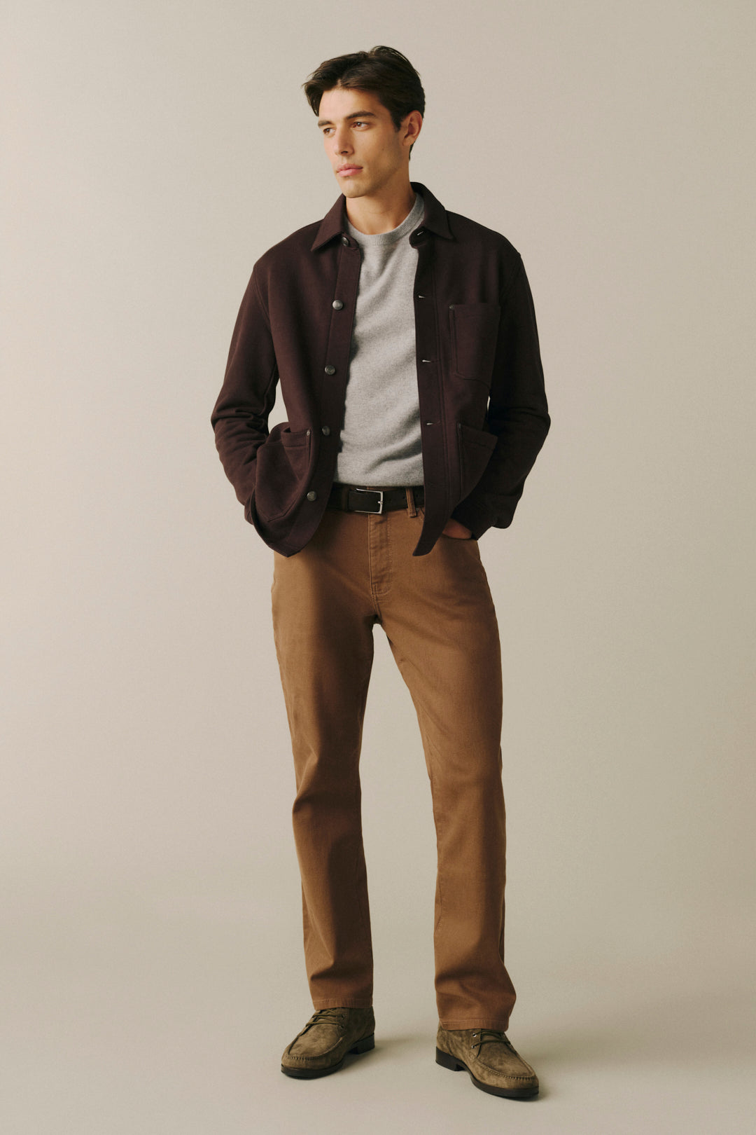 A man modeling a dark brown overshirt over a light gray sweater, paired with brown trousers and brown shoes, standing against a plain beige studio background.