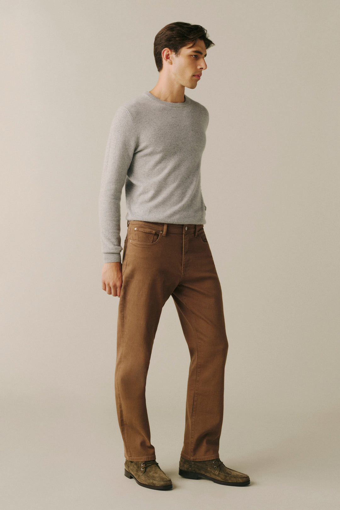 A man wearing a light gray long-sleeve crewneck sweater, brown straight-leg jeans, and brown boots, standing in profile against a plain light beige studio background.