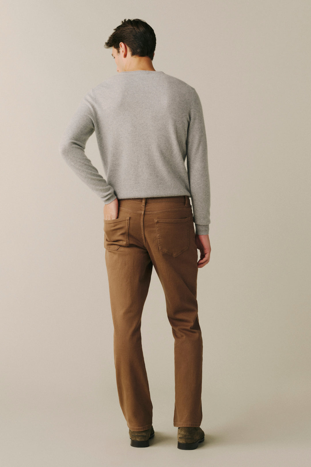 A man shown from behind wearing a light gray long-sleeve sweater and brown trousers, standing against a plain beige studio background.