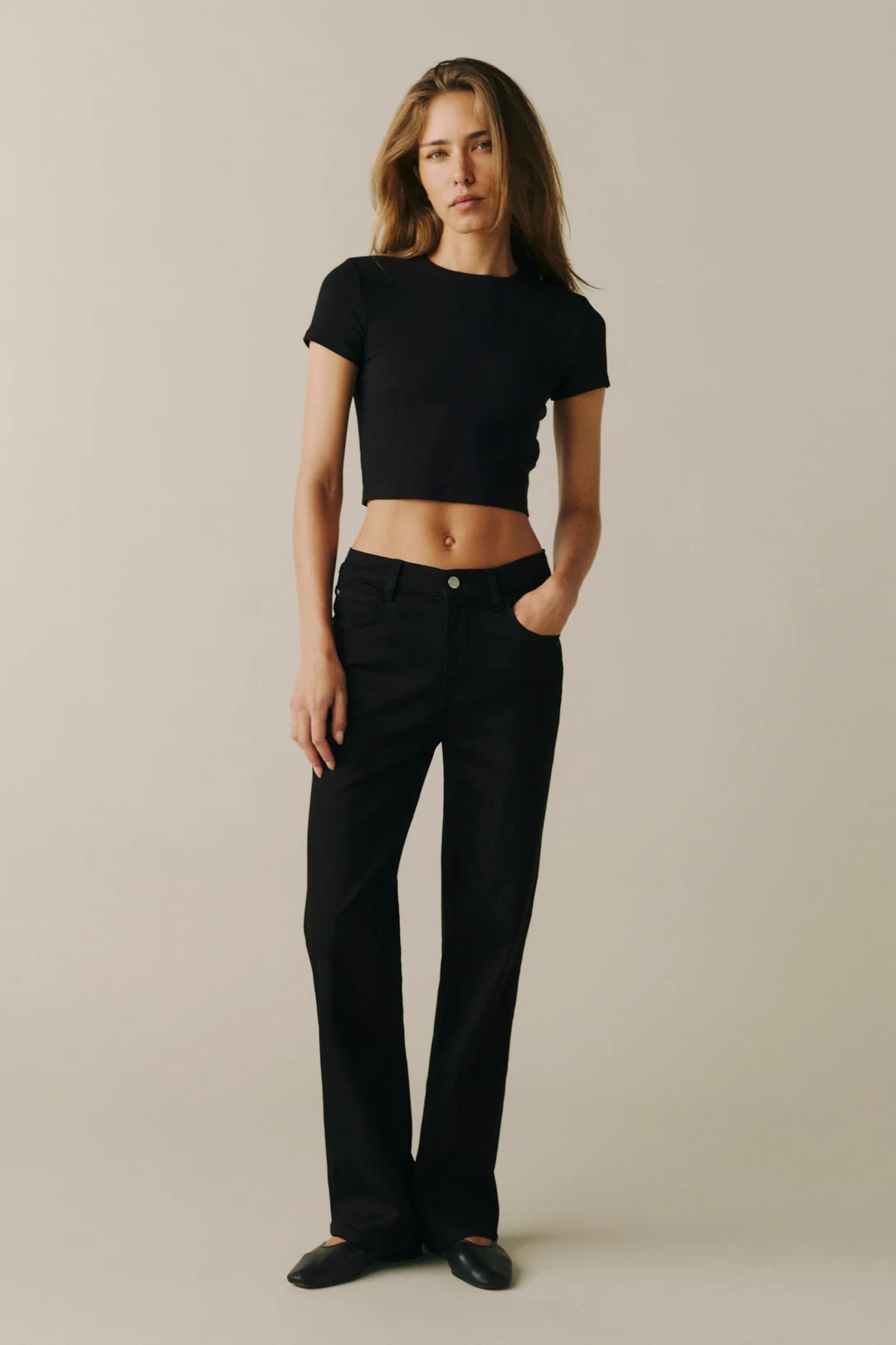 A woman modeling a black short-sleeve crop top and black straight-leg jeans against a plain beige studio background.