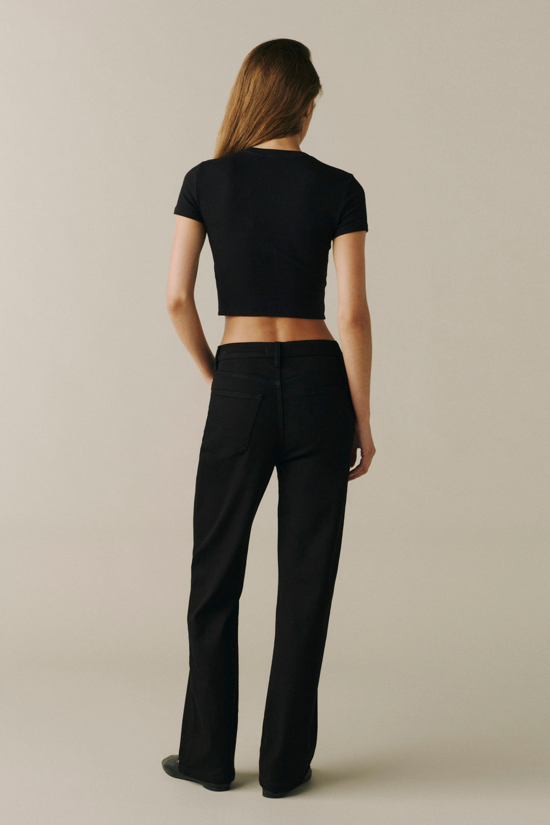 A woman is shown from the back wearing a black short-sleeve crop top and black wide-leg trousers, standing against a plain beige studio background.